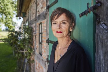 Claudia Wuttke lehnt gegen eine grüne Schuppentür und schaut seitlich lächelnd in die Kamera