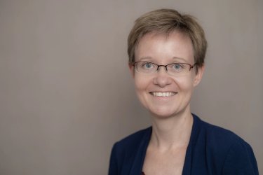 Meike Blatzheim steht vor einer beigen Wand und lächelt in die Kamera