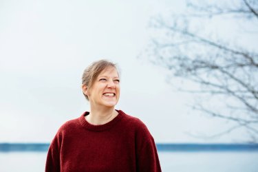 Petra Hucke steht vor einem See im roten Pullover und schaut lachend in die Ferne